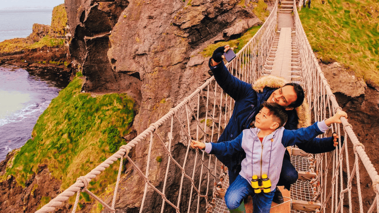 An adult and child pause on Carrick-a-Rede Rope Bridge to take a selfie, with rugged coastal cliffs, grassy headlands and the sea stretching out below.